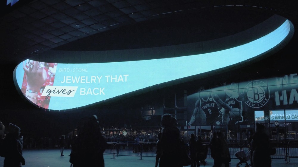 Large curved digital display showing jewelry advertisement in modern sports arena with silhouetted visitors below